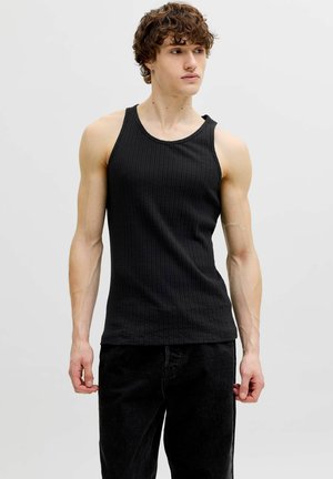 Jeune homme aux cheveux bouclés portant un débardeur noir côtelé et un pantalon noir, debout devant un fond clair uni.