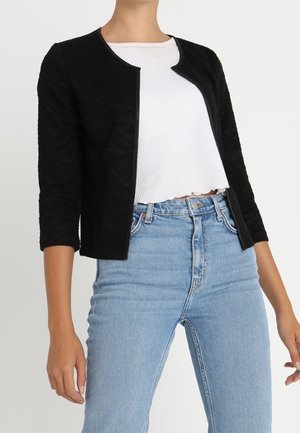 Kvinde iført en sort cardigan med åben front, hvid cropped top og højtaljede lyseblå jeans, stående mod en ensfarvet baggrund.