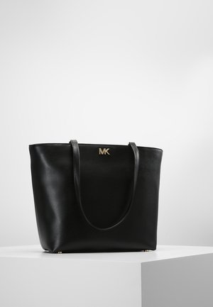 Sort læder tote taske med to skulderstropper og guld MK logo foran, placeret på en hvid piedestal mod en ensfarvet baggrund.