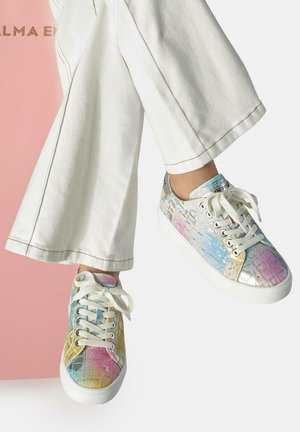 Pantalones blancos de piernas anchas combinados con zapatillas de cuero multicolor en tonos pastel con cordones blancos, llevados por una persona sentada contra un fondo rosa y blanco.