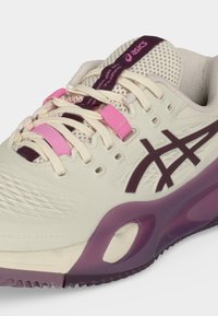 Κοντινή λήψη μπεζ παπουτσιού τρεξίματος Asics με μοβ λογότυπο, ροζ θηλιές κορδονιών και μοβ λεπτομέρειες στη σόλα.