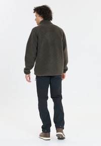 Fleecejacka i mörk oliv med hög krage, med en mjuk textur och ribbade ärmmuddar, tillsammans med svarta tekniska byxor och bruna stövlar.