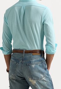 Muž v světle modré košili s dlouhým rukávem, s vyhrnutými rukávy, hnědým opaskem a vybledlými modrými džínami s opotřebovanými místy, zobrazený zezadu.