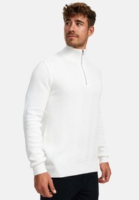 Mann iført hvit teksturert genser med halvglidelås og høy krage, stående og ser til siden mot en ensfarget bakgrunn.