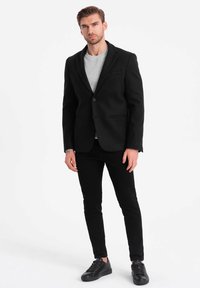 Zwarte blazer met notch-lapels, enkelknopenluiting en twee voorzakken, gecombineerd met een grijs sweater en zwarte broek. Zwarte sneakers.