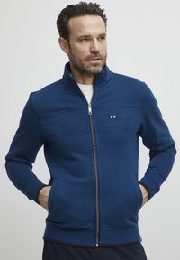 Mørkeblå hettejakke med glidelås og høy krage, teksturerte detaljer, to sidelommer og en liten logo på brystet. Mykt stoff.