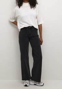 Jeans neri a vita alta e a gamba larga abbinati a un top bianco a maniche corte con collo alto e sneakers grigie, che mostrano un fit rilassato e uno stile casual.