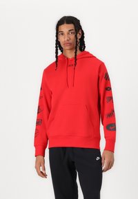 Sarkans Nike kapuci džemperis ar kangaroo kabatu, aukliņu kapuci un melniem grafiskiem ornamentiem uz piedurknēm. Apvienots ar melnām biksēm.