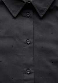 Zwarte button-up shirt met een gestructureerde kraag, een rij zwarte knopen en kleine decoratieve studs verspreid over de stof.