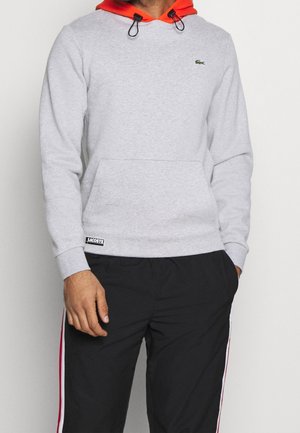 Mand iført lysegrå Lacoste hættetrøje med orange hætte og sorte bukser med røde og hvide side striber, hænder i lommerne.