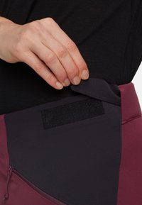 Pantaloni a vita alta in bordeaux e viola scuro, con chiusura in velcro e una tasca laterale con coulisse. Tessuto dalla texture liscia.