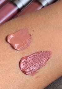 Två prover av krämiga mauve-rosa läppstiftsnyanser applicerade på mediumbrun hud nära bifogade läppglansrör.