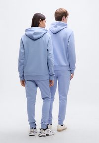 Ανοιχτό μπλε φούτερ με κουκούλα και αντίστοιχο παντελόνι φόρμας από μαλακό υλικό. Διαθέτει ριμπ τελειώματα στους καρπούς και άνετη εφαρμογή. Ορατά αθλητικά παπούτσια στα πόδια.