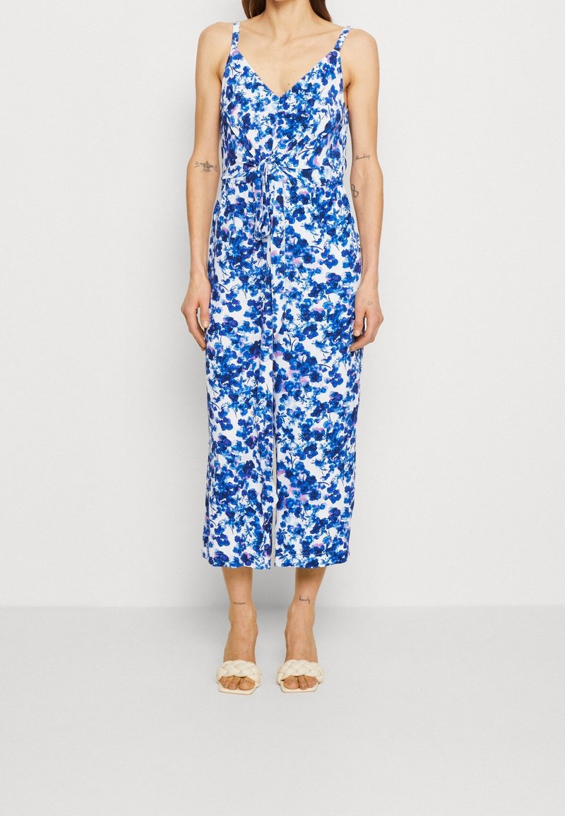 Femme portant une combinaison sans manches à motifs floraux bleus et blancs avec une ceinture nouée à la taille et des sandales blanches tressées à bout ouvert, debout devant un fond uni.