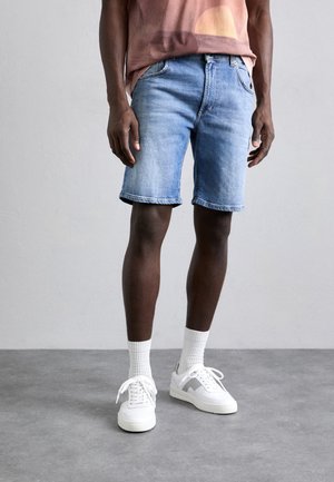 Shorts en denim bleu clair, présentant une coupe droite, un ourlet retroussé et des poches avant. Portés avec des baskets blanches et des chaussettes côtelées.