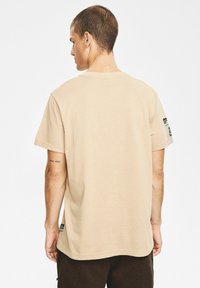 Beżowa bawełniana koszulka z okrągłym dekoltem, krótkimi rękawami i prostym krojem. Posiada małą metkę na dolnym boku oraz napisy na rękawie.