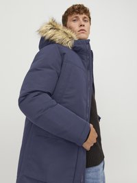 Parka blu navy con cappuccio bordato in pelliccia, chiusura con zip, tasche laterali e texture trapuntata, indossata sopra un maglione scuro e jeans.