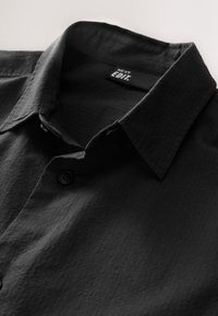 Chemise noire boutonnée avec un tissu texturé, col pointu, et une fermeture à un seul bouton. L'étiquette indique "NEXT EDIT."