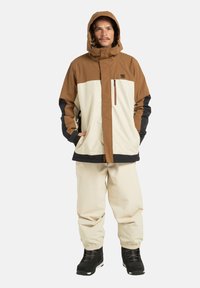 Giacca isolata marrone e crema con cappuccio, maniche nere a contrasto, e pantaloni crema, indossati con stivali da neve neri.