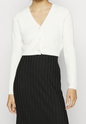 Femme portant un cardigan court blanc à manches longues avec des boutons et une jupe noire côtelée taille haute.