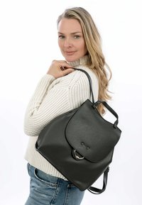 Mochila de cuero negro con un acabado texturizado, solapa plegable, acento de hardware redondo y asas dobles. Lleva sobre el hombro contra un fondo blanco.
