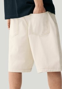 Beige Baumwollshorts in lockerer Passform mit zwei Gesäßtaschen und sichtbaren Nähdetails. Der Saum ist gekrempelt für einen sauberen Abschluss.