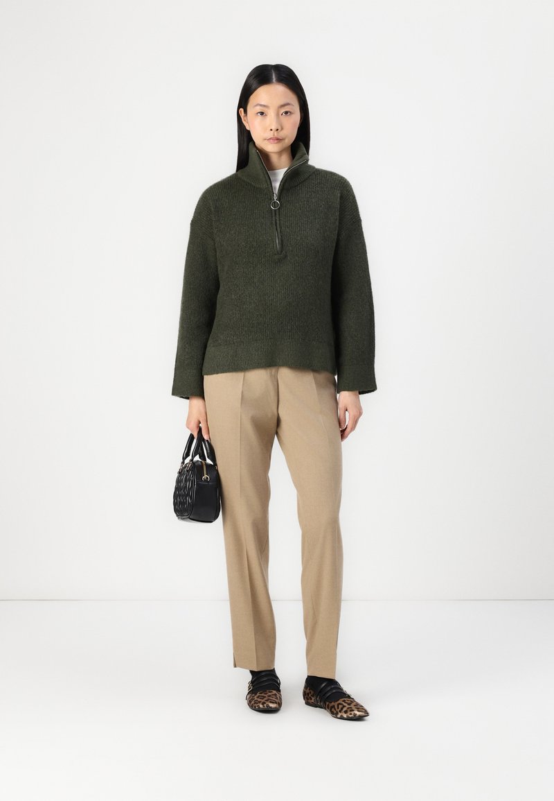 Dunkelgrüner gestrickter Pullover mit einem halben Reißverschlusskragen, kombiniert mit beigen Hosen. Accessoires sind eine kleine schwarze Handtasche und Schuhe mit Leopardenmuster.