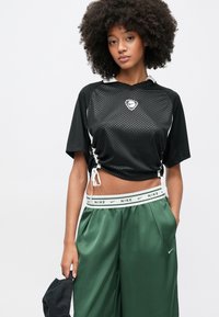 Νέα γυναίκα με σγουρά μαλλιά που φοράει μαύρο κοντό αθλητικό τοπ και πράσινο παντελόνι με φαρδιά μπατζάκια, κρατώντας ένα μαύρο καπέλο στο αριστερό της χέρι.