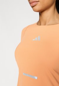 Sportinė palaidinė persikų spalvos, pagaminta iš tekstūruotos medžiagos. Sidabriniai akcentai matomi juostoje ir logotipe, apvalus kaklo iškirpimas ir trumpomis rankovėmis.