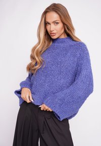 Chunky blue sweater with a high neckline, wide sleeves, and textured knit. Paired with black pleated trousers.