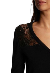 Pull noir à manches longues, avec un col en V et un détail en dentelle sur l'épaule. Texture lisse avec un motif de dentelle complexe.