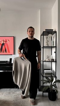 Hellbeiger Pullover mit einem gerippten Rundhalsausschnitt, gehalten an einem Bügel, getragen von einer Person in einem schwarzen T-Shirt und dunkler Hose. Minimalistischer Hintergrund.