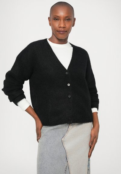 Cardigã preto e felpudo com decote em v e seis botões, combinado com uma camisa branca por baixo. Modelo veste uma saia assimétrica de denim claro.