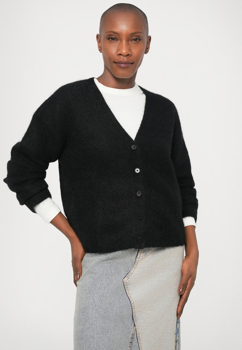 Cardigan nero e morbido con scollo a V e sei bottoni, abbinato a una camicia bianca sotto. Il modello indossa una gonna asimmetrica in denim chiaro.