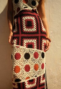 Häkelkleid mit geometrischen Quadraten und Kreisen in Rot, Schwarz und Orange, verziert mit einem strukturierten weißen Spitzenbesatz und offener Stricktechnik.