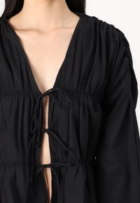 Fekete blúz mély V-nyakkal, redőzött anyaggal, derékra húzható zsinórral és hosszú ujjakkal. Sima textúra, könnyű anyag.
