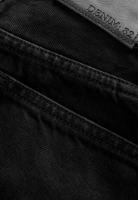 Černá denimová tkanina s viditelnými švy a koženou etiketu s nápisem "DENIM: 82" vyraženým písmem. Uzavřený pohled.