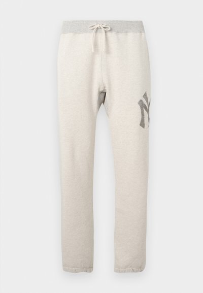 Pantaloni de trening bej deschis, cu un brâu elastic gri, șnur, buzunare laterale și un grafic gri închis pe cosul stâng.