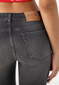 Jean en denim gris foncé avec un design ajusté. Les caractéristiques incluent une étiquette en cuir, des poches arrière cousues et une texture lisse.