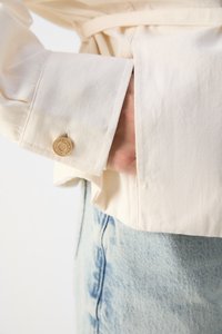 Giacca in tessuto beige con tasca frontale e polsini con bottoni. Il materiale sembra liscio con una lieve texture. Indossata sopra un jeans azzurro chiaro.