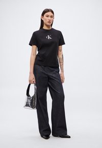 Svart bomulls-t-skjorte med sølvlogo, kombinert med mørk denim vidbukser. Skinnlignende svarte loafers og en gjennomsiktig veske med håndtak.