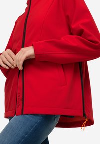 Giacca leggera rossa con chiusura a zip, tasche laterali e un accento arancione sulla zip. Texture liscia e raffinata.
