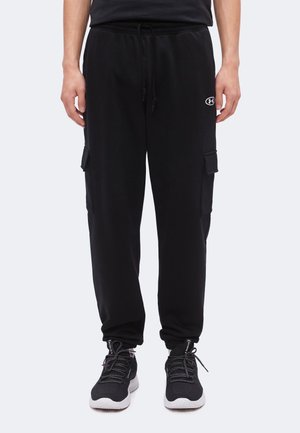 Mand iført sorte cargo sweatpants med snøre i taljen og sorte træningssko på en lys baggrund.