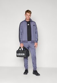 Lila melegítőfelső fekete csíkokkal, fekete póló és fekete táska fehér "adidas" logóval. Textúrált anyag és testhezálló fazon.