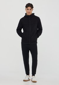 Felpa nera da uomo con zip e colletto alto, abbinata a pantaloni joggers coordinati. Tessuto: morbido, texturizzato. Scarpe bianche con accenti neri.