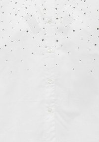 Balta marškiniai su lygiu audiniu ir išsibarsčiusiais cirkonių papuošimais. Klasikinis marškinių stilius su penkiais matomais mygtukais.