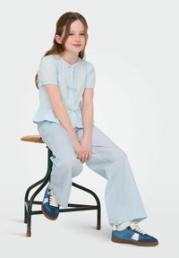 Haut bleu clair à manches courtes avec détail noué, associé à un pantalon large bleu clair et des baskets bleues avec accents blancs, assis sur un tabouret.