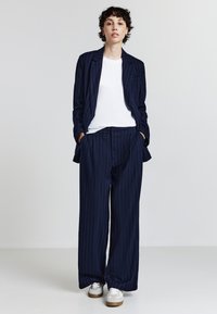 Marineblau gestreifter Anzug mit weit geschnittenen Hosen und einem maßgeschneiderten Blazer, kombiniert mit einem weißen T-Shirt und hellen Sneakern. Glatte Textur, lässige Passform.