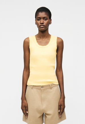 Woman with short hair wearing a sleeveless yellow top and beige shorts, standing against a plain light background.