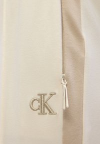 Μπουφάν Calvin Klein με υφαντό σε ανοιχτό μπεζ χρώμα, διαθέτει φερμουάρ, κεντημένο λογότυπο "cK" με μεταλλική κλωστή, ομαλή υφή και μινιμαλιστικό σχεδιασμό.